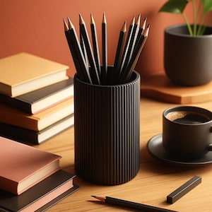 Op de afbeelding: Een zwarte pennenhouder gevuld met geslepen potloden, een kopje koffie en een stapel boeken op een houten oppervlak. De potloden hebben een zwarte behuizing en een gouden punt. De koffiekop en schotel zijn zwart. De boeken hebben verschillend gekleurde kaften.