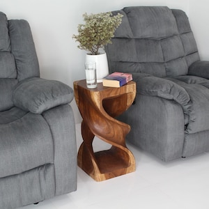 May include: Two gray reclining chairs with a wooden side table between them. The table is carved in a spiral shape and has a book on top. A small plant is in a white vase on the table.