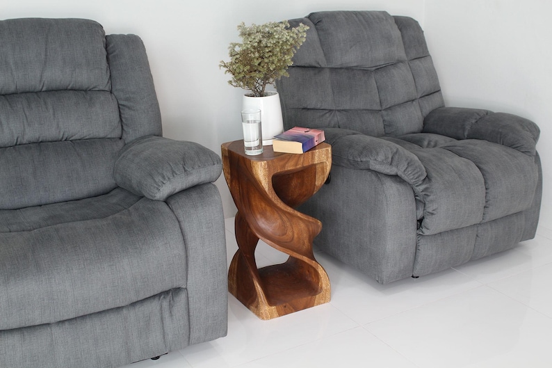 May include: Two gray reclining chairs with a wooden side table between them. The table is carved in a spiral shape and has a book on top. A small plant is in a white vase on the table.