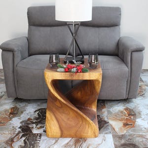 May include: A wooden side table with a unique twisted design. The table is a warm brown color and has a smooth finish. Two glasses and a decorative floral arrangement sit on top. A gray sofa and lamp are in the background.