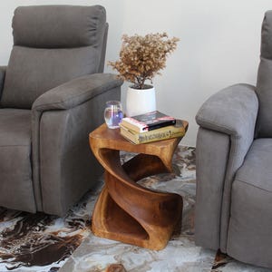 May include: A wooden side table with a unique twisted design. The table is made of natural wood and has a rustic finish. It is placed between two gray armchairs in a living room setting.