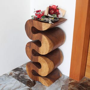 May include: A wooden side table with a unique, wavy design. The table is made of dark brown wood and has three tiers. A small tray sits on the top tier, holding a bouquet of red flowers.