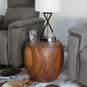 May include: A solid, wooden side table with a natural finish. Two clear glasses of water sit on top. A black lamp with a white lampshade is behind the glasses. Two gray upholstered recliners are on either side of the table.