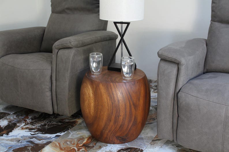May include: A solid, wooden side table with a natural finish. Two clear glasses of water sit on top. A black lamp with a white lampshade is behind the glasses. Two gray upholstered recliners are on either side of the table.