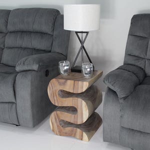 May include: A wooden side table with a unique, wavy design. The table is made of natural wood and has a light brown color. Two gray armchairs are in the background.
