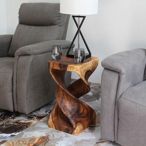 May include: A wooden side table with a unique twisted design. The table is made of dark brown wood and has a natural finish. It is sitting between two gray armchairs in a living room.