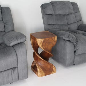 May include: A wooden end table with a unique twisted design. The table is made of dark brown wood and has a natural finish. It is sitting in front of two gray reclining chairs.