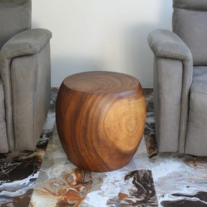 May include: A solid wood side table with a natural finish, positioned between two gray upholstered armchairs. The table has a rounded, barrel-like shape and sits on a patterned tile floor. The wood grain is visible.