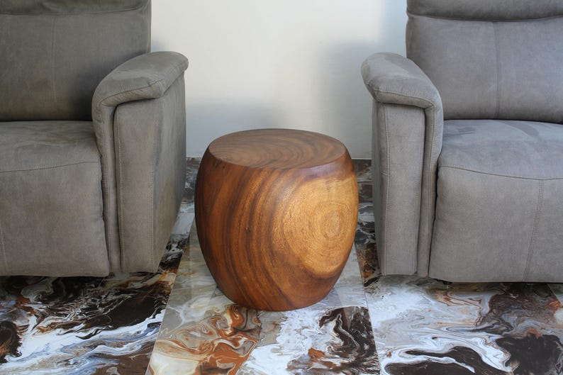 May include: A solid wood side table with a natural finish, positioned between two gray upholstered armchairs. The table has a rounded, barrel-like shape and sits on a patterned tile floor. The wood grain is visible.