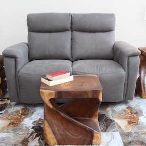 May include: A gray fabric loveseat with two wooden side tables and a wooden coffee table. The coffee table is a unique design with a twisted shape. The loveseat is in a living room setting with a white wall and a brown tiled floor.