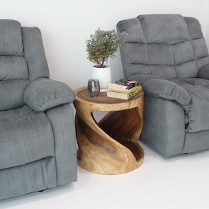 May include: Two gray fabric recliners with a wooden side table between them. The table has a twisted design and is decorated with a potted plant and books.