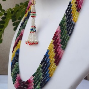 May include: A colourful multi-strand necklace with beads in shades of pink, red, yellow, green, and blue. A white beaded tassel with red, green, and blue accents hangs from the top. The necklace is displayed on a white bust.