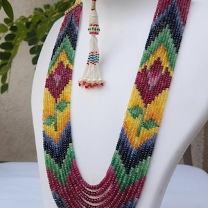 Op de afbeelding: Een meerstrengs kralenketting met een bloemmotief. De ketting heeft een kleurenpalet van rode, gele, groene en blauwe kralen. Een kralenkwast hangt aan de ketting. De ketting wordt getoond op een witte mannequin.