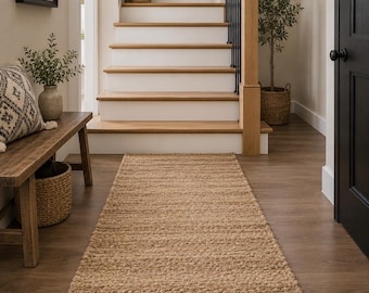 Puede incluir: Una alfombra de pasillo rectangular hecha de fibras de yute tejidas en color tostado natural con una construcción trenzada gruesa y texturizada. La alfombra se extiende sobre un suelo de madera, con un patrón de tejido denso y uniforme.