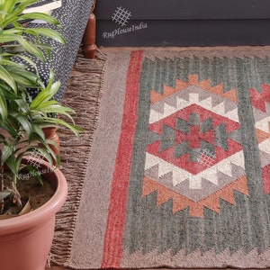 May include: A green, red, and white patterned rug with fringe on the edges. The rug has a geometric design and is made of wool. The rug is on a wooden floor and there is a plant in a pot in the foreground. The text 'RugHouseIndia' is visible in the background.