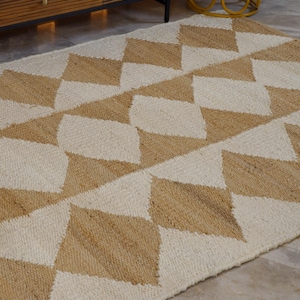 Può includere: Un tappeto rettangolare con un motivo geometrico a forma di rombi beige e bianco sporco alternati. Il tappeto è realizzato in un materiale tessuto ed è posizionato su un pavimento di colore chiaro. È visibile una sedia di legno con un cuscino fantasia.