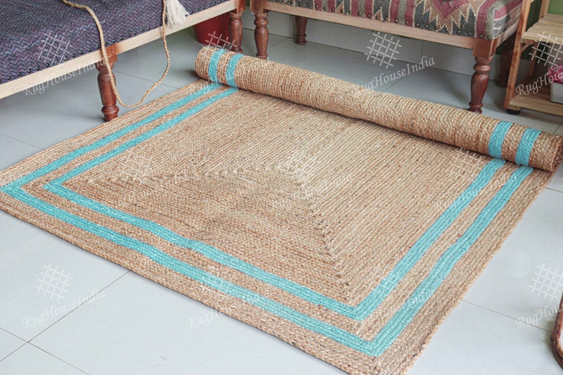 May include: A natural fiber rug with a square shape and a light brown color. The rug has a blue border with two stripes around the edges.