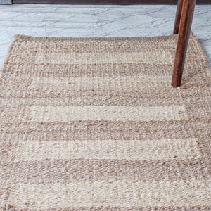 May include: A rectangular, woven rug with alternating beige and off-white horizontal stripes. The rug is made of a natural fiber and has a textured appearance. A wooden chair leg is partially visible, resting on the rug.
