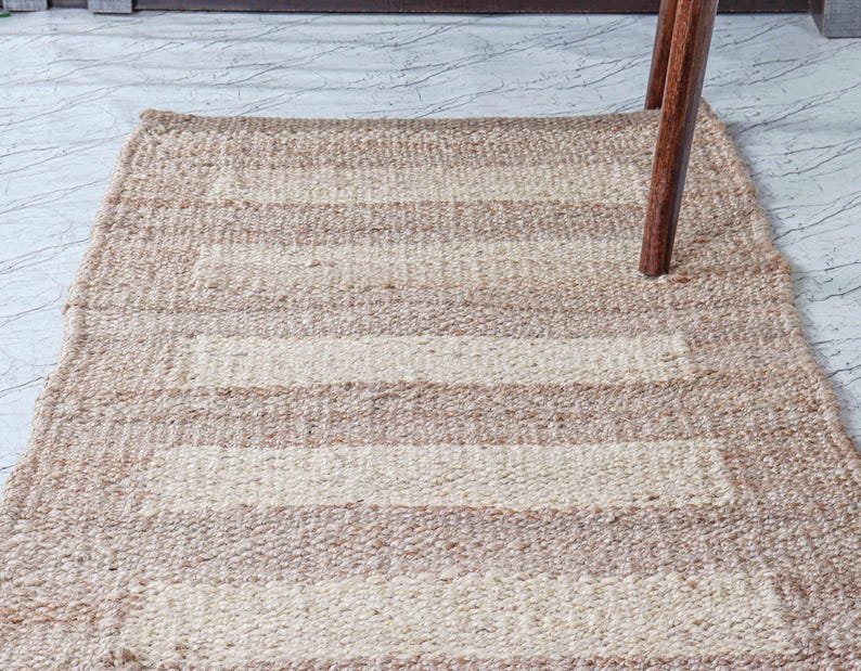 May include: A rectangular, woven rug with alternating beige and off-white horizontal stripes. The rug is made of a natural fiber and has a textured appearance. A wooden chair leg is partially visible, resting on the rug.