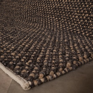 May include: Close-up of a textured rug with dark brown and beige fibres. The rug has a woven appearance with a visible pattern and a white border. The rug is made of a soft material.