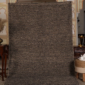 May include: A large, rectangular rug with a textured, woven design in shades of brown and beige. The rug has a thick pile and a white border along the bottom edge. The rug is displayed against a decorative wall.