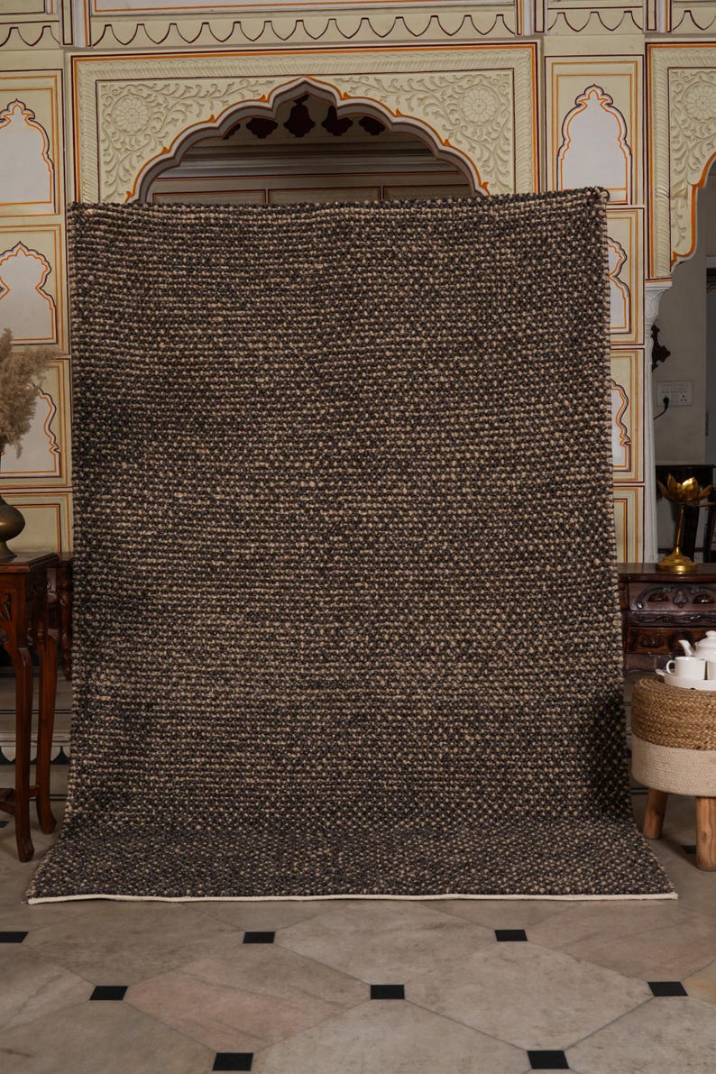 May include: A large, rectangular rug with a textured, woven design in shades of brown and beige. The rug has a thick pile and a white border along the bottom edge. The rug is displayed against a decorative wall.
