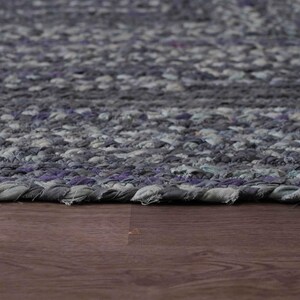 May include: Close-up of a braided rug with a mix of gray and purple tones. The rug's texture is visible, with a detailed pattern. The rug is placed on a dark brown wooden surface.