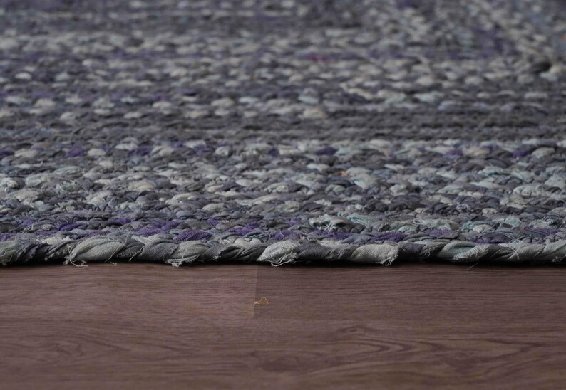 May include: Close-up of a braided rug with a mix of gray and purple tones. The rug's texture is visible, with a detailed pattern. The rug is placed on a dark brown wooden surface.
