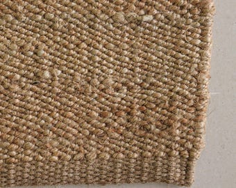 May include: Close-up of a natural fibre rug with a woven texture. The rug is a warm, neutral brown colour, with variations in tone. The weave pattern is visible, creating a textured surface. The edge of the rug is visible, showing a finished border.