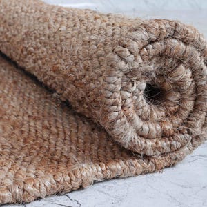 May include: Rolled-up jute rug with a natural brown color and a woven texture. The rug's edge is visible, showcasing the tightly woven fibers. The rolled section reveals the rug's thickness and construction, suitable for home decor.