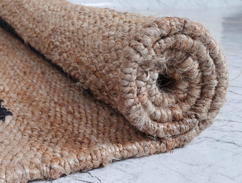 May include: Rolled-up jute rug with a natural brown color and a woven texture. The rug's edge is visible, showcasing the tightly woven fibers. The rolled section reveals the rug's thickness and construction, suitable for home decor.