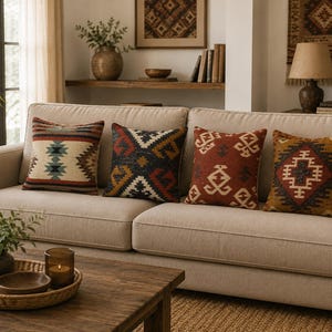 May include: Four square throw pillows arranged on a beige sofa. Each pillow features a distinct woven pattern with geometric shapes in shades of red, brown, cream, black, and blue. The fabric appears textured with a visible weave throughout the designs.