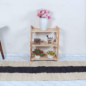 Op de afbeelding: Een houten plank met drie niveaus en decoratieve items, waaronder een vaas met roze bloemen, staat tegen een witte muur. Een beige tapijt met een zwarte streep ligt op de vloer. Links een kleine witte tafel met houten poten.