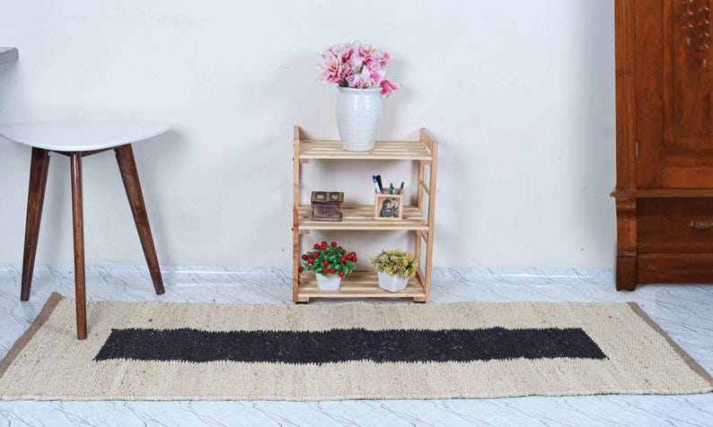 Op de afbeelding: Een houten plank met drie niveaus en decoratieve items, waaronder een vaas met roze bloemen, staat tegen een witte muur. Een beige tapijt met een zwarte streep ligt op de vloer. Links een kleine witte tafel met houten poten.