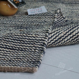 May include: A rectangular area rug featuring a thick, woven texture with alternating horizontal stripes of dark navy blue and natural beige fibers. The rug has a visible, sturdy braided construction with a solid brown border along the bottom edge.