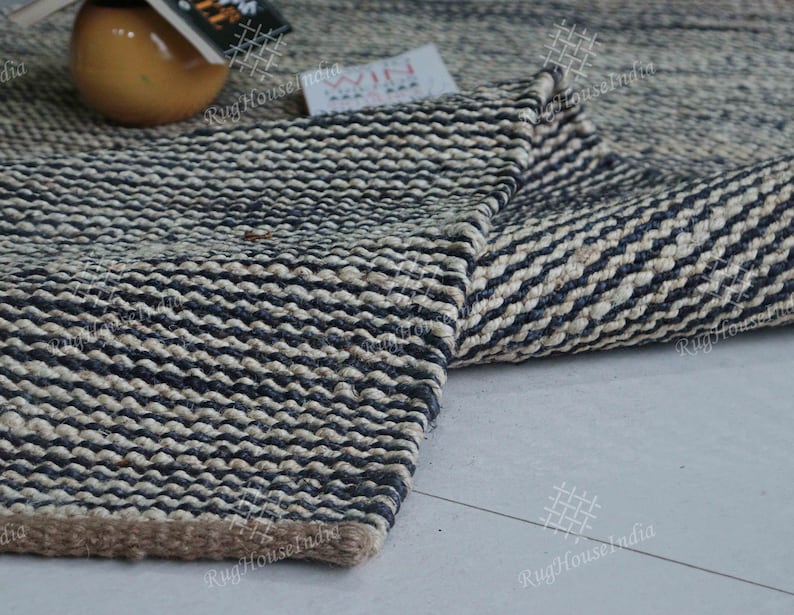 May include: A rectangular area rug featuring a thick, woven texture with alternating horizontal stripes of dark navy blue and natural beige fibers. The rug has a visible, sturdy braided construction with a solid brown border along the bottom edge.
