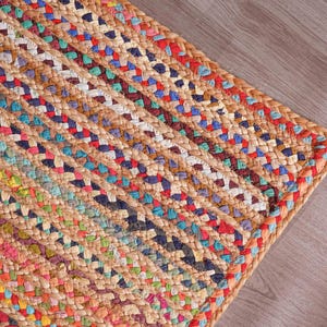 Puede incluir: Alfombra de yute trenzado con coloridos detalles tejidos. La alfombra presenta una base de color beige natural con rayas rojas, azules, verdes y blancas. Se ve la esquina de la alfombra, mostrando la construcción trenzada y la textura de las fibras.