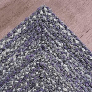 May include: Braided rug corner in shades of gray, white, and purple. The rug's texture is visible, with a mix of colors creating a textured pattern. The rug is placed on a wooden surface.
