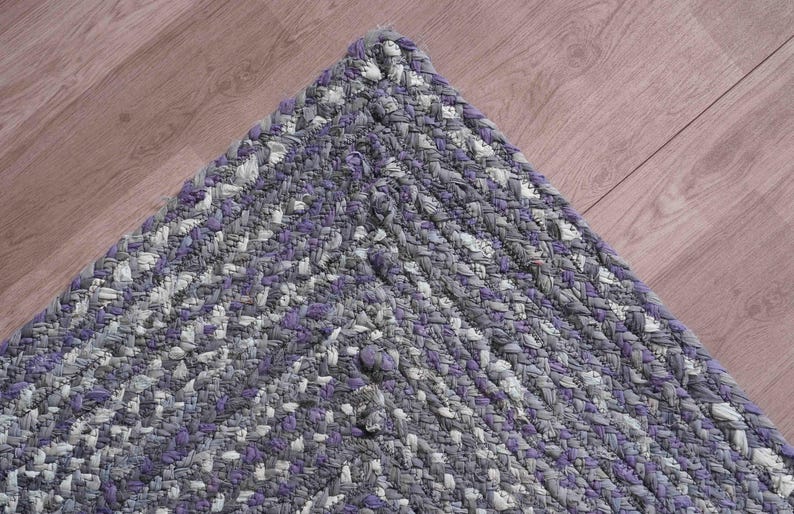 May include: Braided rug corner in shades of gray, white, and purple. The rug's texture is visible, with a mix of colors creating a textured pattern. The rug is placed on a wooden surface.