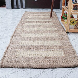 May include: A rectangular, beige and off-white striped jute rug. The rug has a woven texture and a border. The stripes are horizontal and run the length of the rug. The rug is on a marble floor.