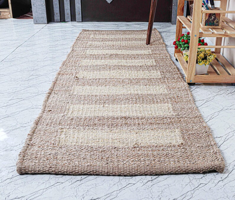 May include: A rectangular, beige and off-white striped jute rug. The rug has a woven texture and a border. The stripes are horizontal and run the length of the rug. The rug is on a marble floor.