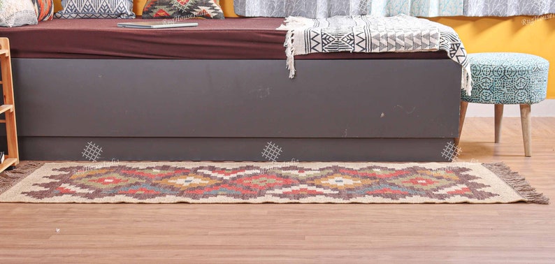 May include: A brown and beige patterned jute rug with fringe on the ends. The rug is laid out on a wooden floor in front of a gray couch and a teal stool.