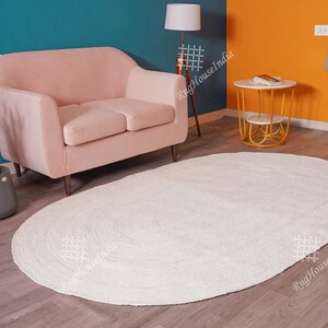 May include: An oval-shaped area rug with a solid off-white color and a braided texture. The rug features a continuous circular pattern created by the braided material, lying flat on a wooden floor surface in front of a light pink sofa.