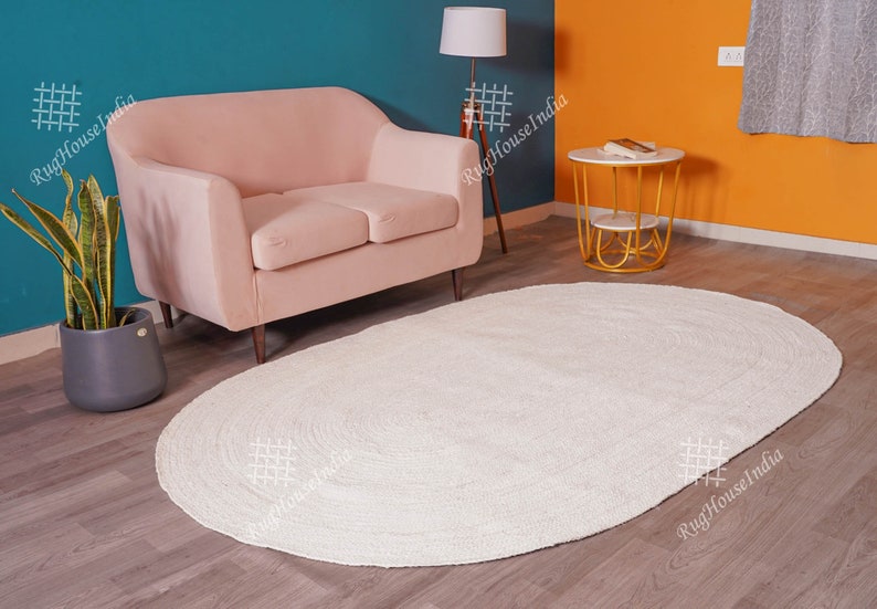 May include: An oval-shaped area rug with a solid off-white color and a braided texture. The rug features a continuous circular pattern created by the braided material, lying flat on a wooden floor surface in front of a light pink sofa.