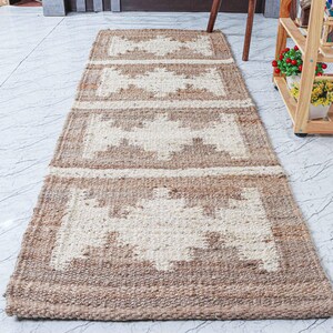 May include: A rectangular jute runner rug with a geometric design. The rug features a repeating pattern of cream-colored geometric shapes framed by a border of brown jute. The rug is placed on a light-colored tile floor.