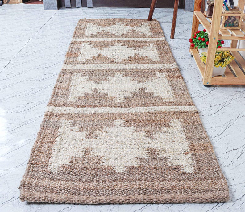 May include: A rectangular jute runner rug with a geometric design. The rug features a repeating pattern of cream-colored geometric shapes framed by a border of brown jute. The rug is placed on a light-colored tile floor.
