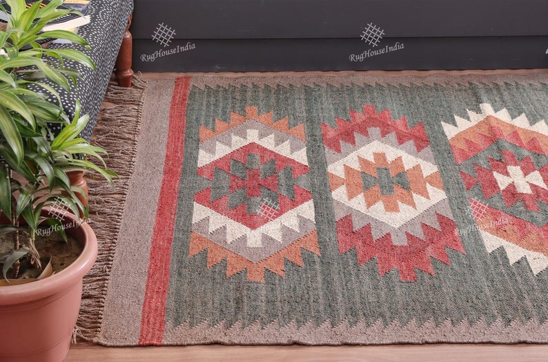 May include: A green, red, and white patterned rug with fringe on the edges. The rug has a geometric design and is made of wool. The rug is on a wooden floor and there is a plant in a pot in the foreground. The text 'RugHouseIndia' is visible in the background.
