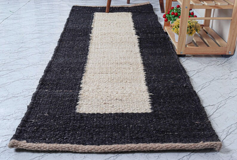 Op de afbeelding: Een rechthoekig jute vloerkleed met een zwarte rand en een cr&egrave;mekleurig middenstuk. Het vloerkleed heeft een geweven textuur en een bruine rand. Dit vloerkleed is geschikt voor een woon- of slaapkamer.
