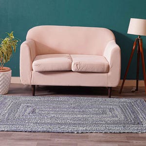 May include: A rectangular, braided rug in shades of gray and white sits on a wooden floor. A light pink loveseat, a potted plant, a wooden tripod lamp, and a small side table with a marble top are in the background.