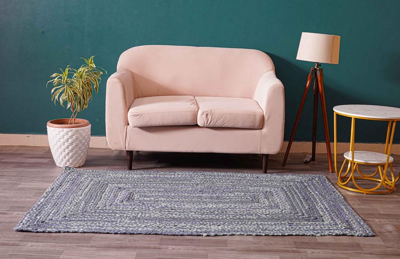 May include: A rectangular, braided rug in shades of gray and white sits on a wooden floor. A light pink loveseat, a potted plant, a wooden tripod lamp, and a small side table with a marble top are in the background.